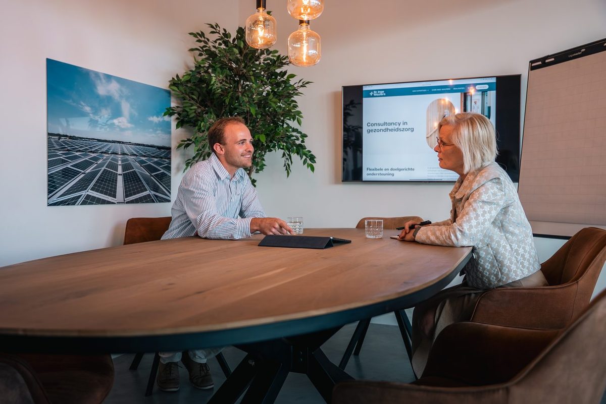 Twee mensen in gesprek aan een ovale vergadertafel, met «Consultancy in gezondheidszorg» op het scherm